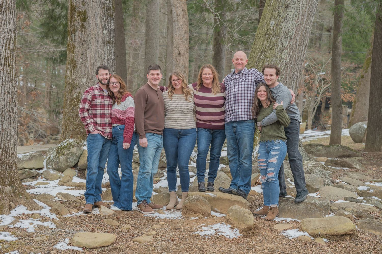 A group of eight people stand together for a photo in a snowy forest setting with large trees and rocks by Proposal Photographer TN.