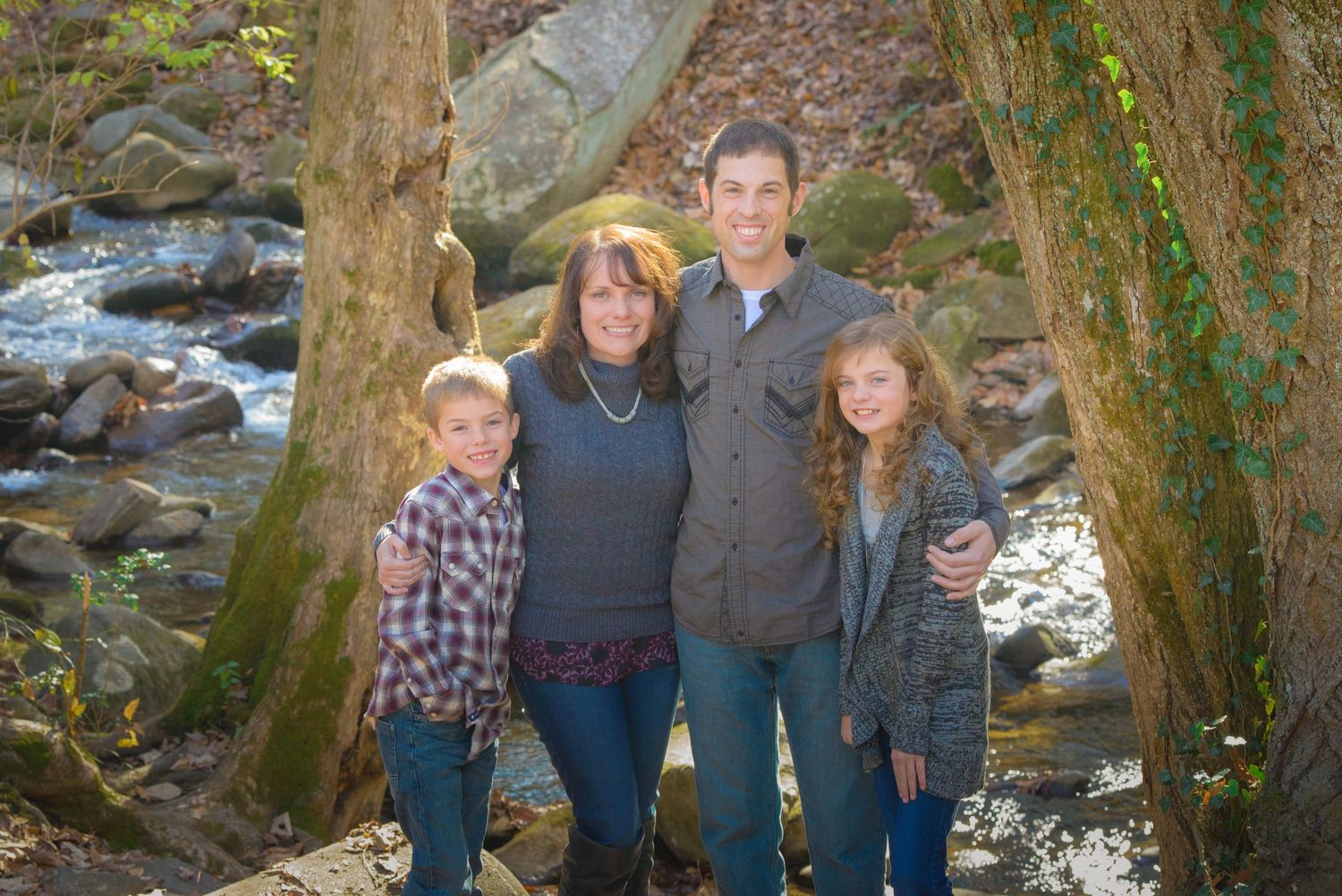 Family poses for a portrait by a scenic creek with rock formations and tree trunks framing the natural outdoor setting in Sevierville TN.