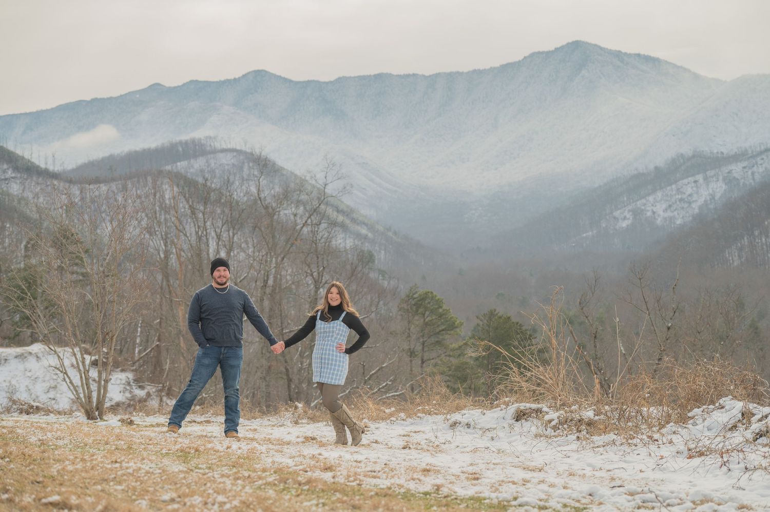 Two people in winter attire leap together while holding hands with misty mountain ranges in the distance on a snowy hillside in Gatlinburg.