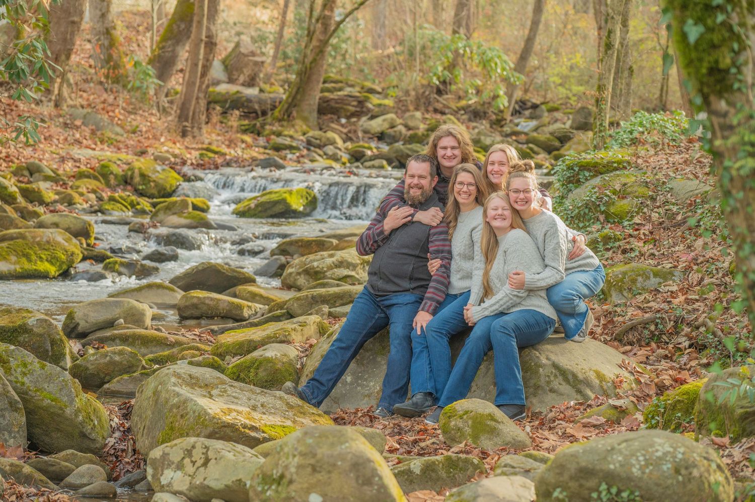 Family sitting together on moss-covered rocks by a flowing stream in autumn woods during outdoor photoshoot by Gatlinburg TN Photographer.