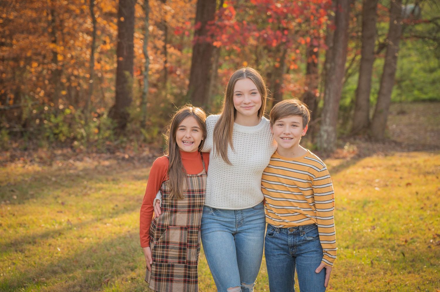 Three kids stand close together outdoors against a backdrop of colorful autumn trees and golden sunlight in Townsend TN.