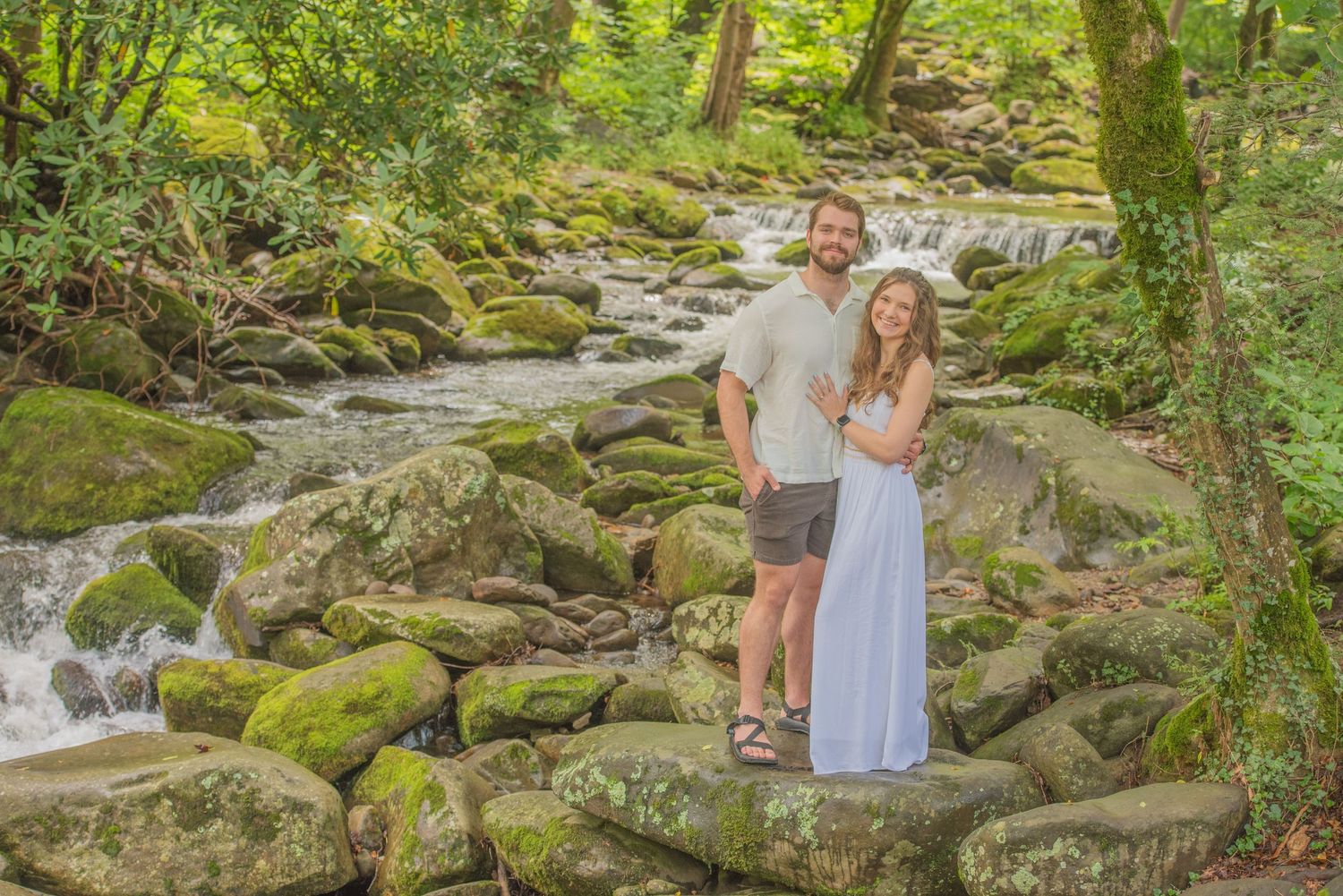 Couple stand on mossy rocks beside a cascading forest stream in a lush green woodland setting by Gatlinburg TN Photographers.