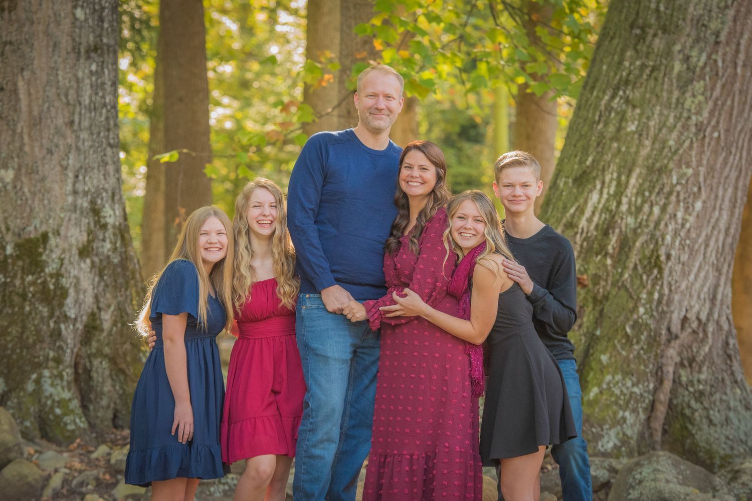 Family group standing together between large tree trunks in a sunlit forest setting during autumn by Pigeon Forge TN Photographer.