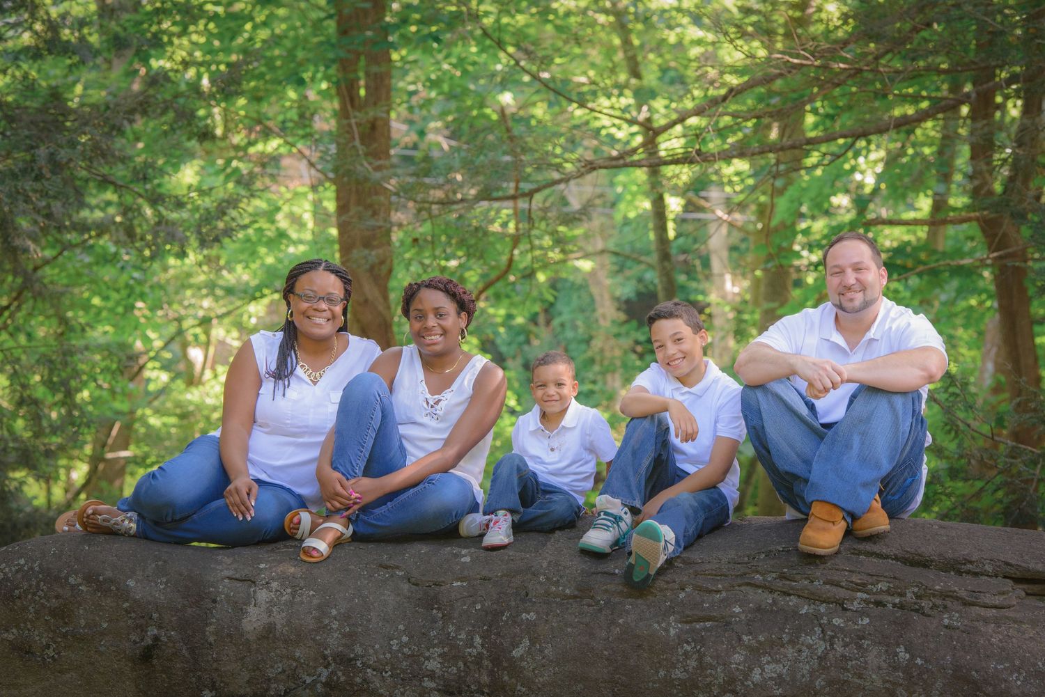A interracial family wearing blue and white clothing sits together on a fallen log in a sunlit forest setting in Townsend TN.