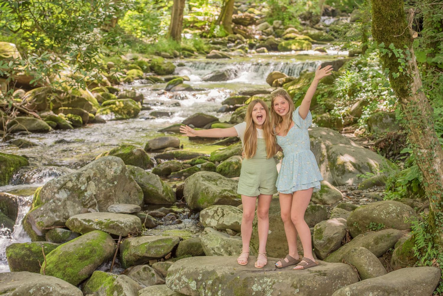 Two sisters in summer outfits stand joyfully on mossy rocks by a cascading forest stream on a sunny day Pigeon Forge TN.