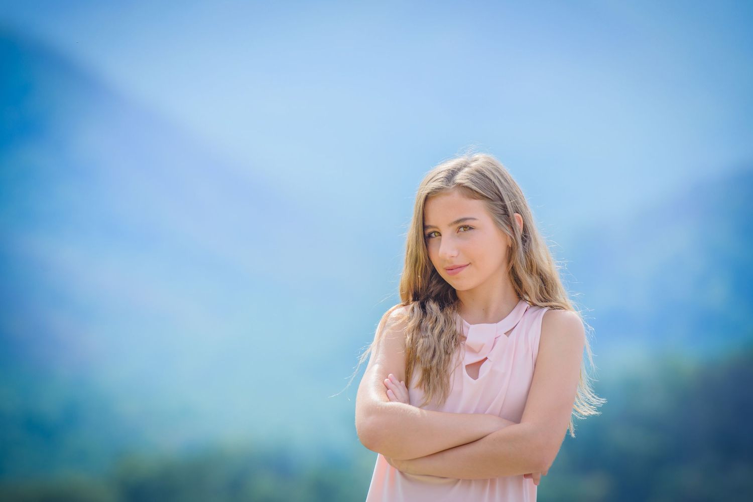 Pigeon Forge Photographer portrait of a young teen in a pink blouse stands with arms crossed against a dreamy blue mountain backdrop.