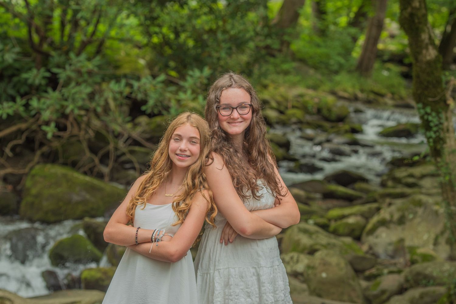 Two sisters pose together by a flowing stream in a lush forest setting wearing light colored dresses in Gatlinburg TN.