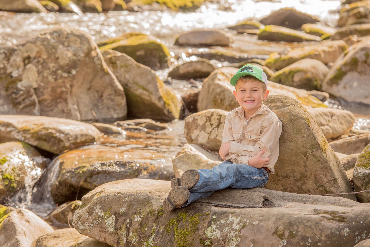 A small figure in a green hat and casual clothing sits on large brown rocks by a stream on a sunny day by Pigeon Forge TN Photographer.