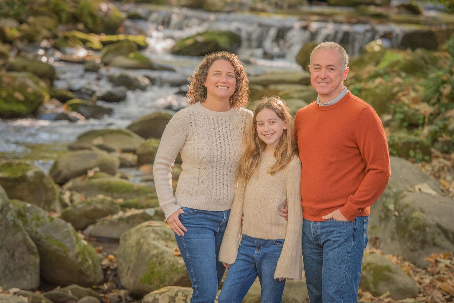 Family in coordinating sweaters pose together on mossy rocks by a flowing stream on a sunny day by Pigeon Forge TN photographers.