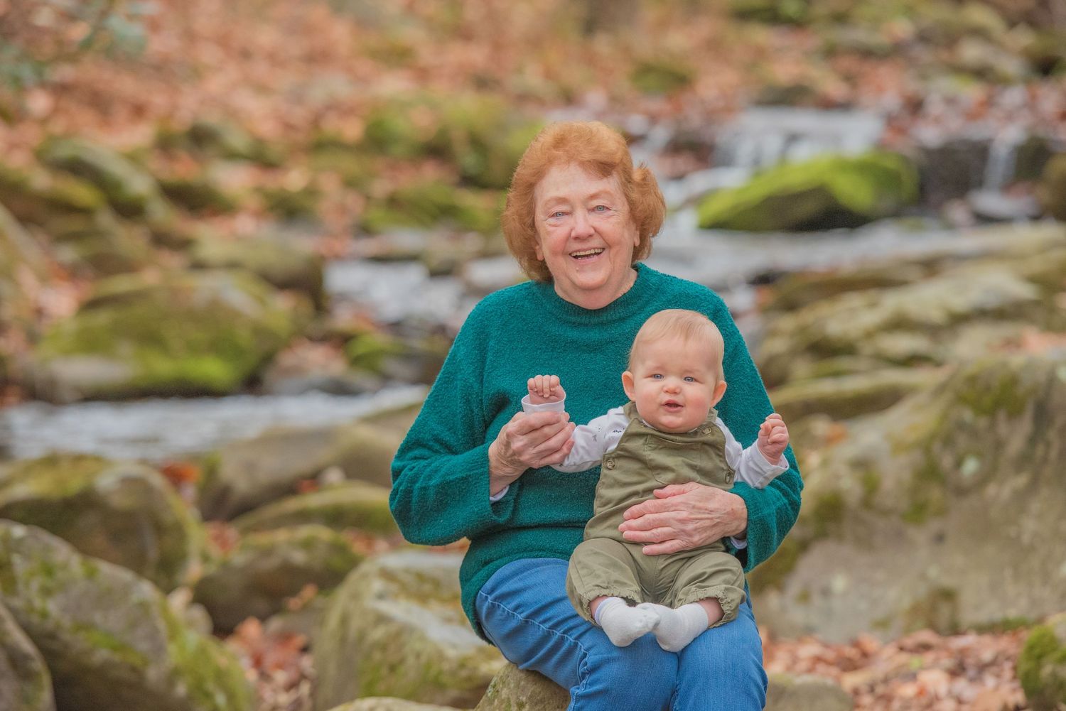 Grandma and baby sit together on rocky terrain with autumn foliage and a stream in the background by Gatlinburg TN Photographer.