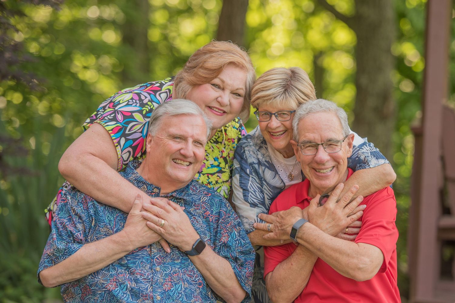 Two senior couples embrace and laugh together in a sunny outdoor setting surrounded by green foliage by Pigeon Forge Photographers.