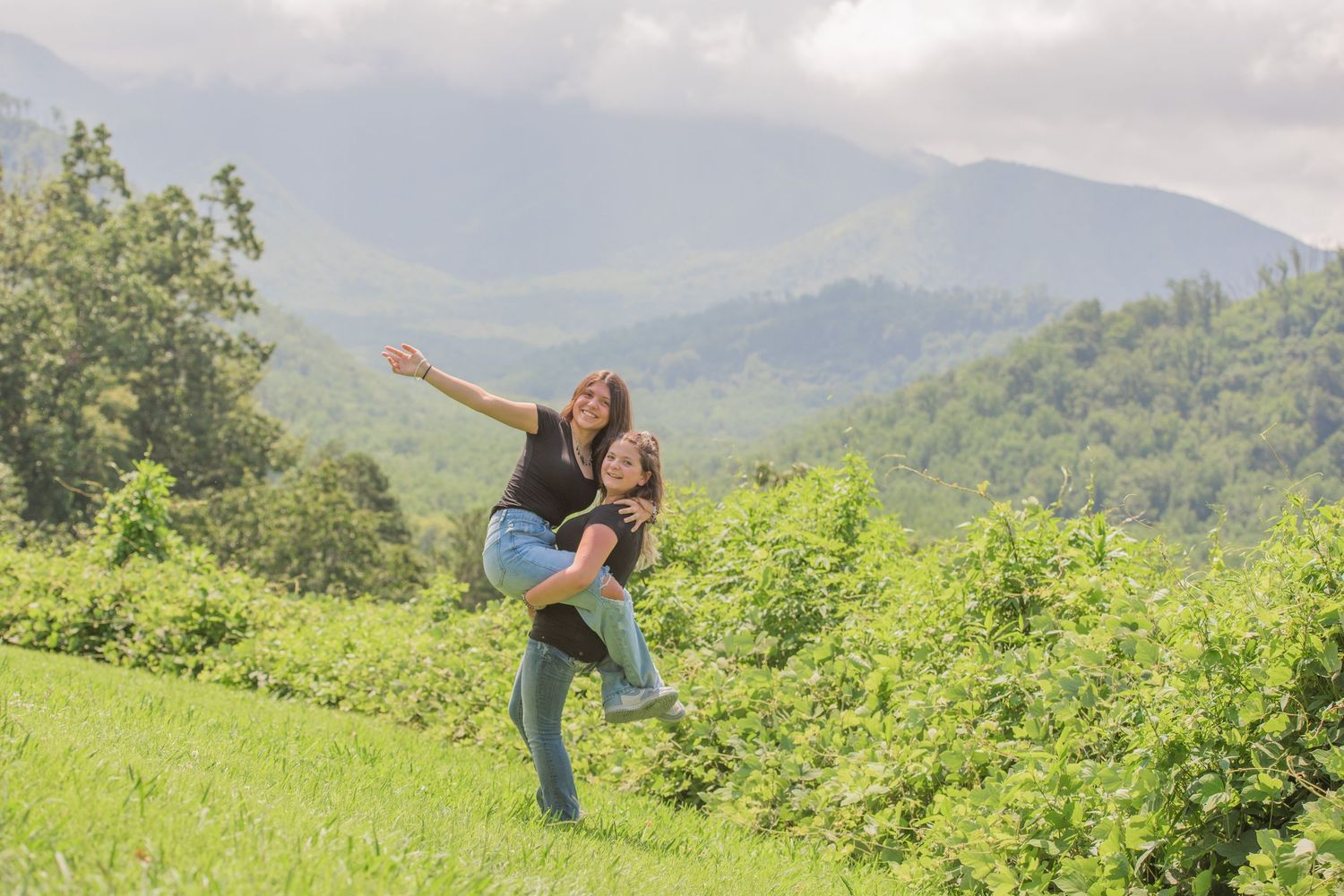 Pigeon Forge Photographers portrait of sisters enjoying a playful piggyback ride on a grassy hill with misty mountains in the background.