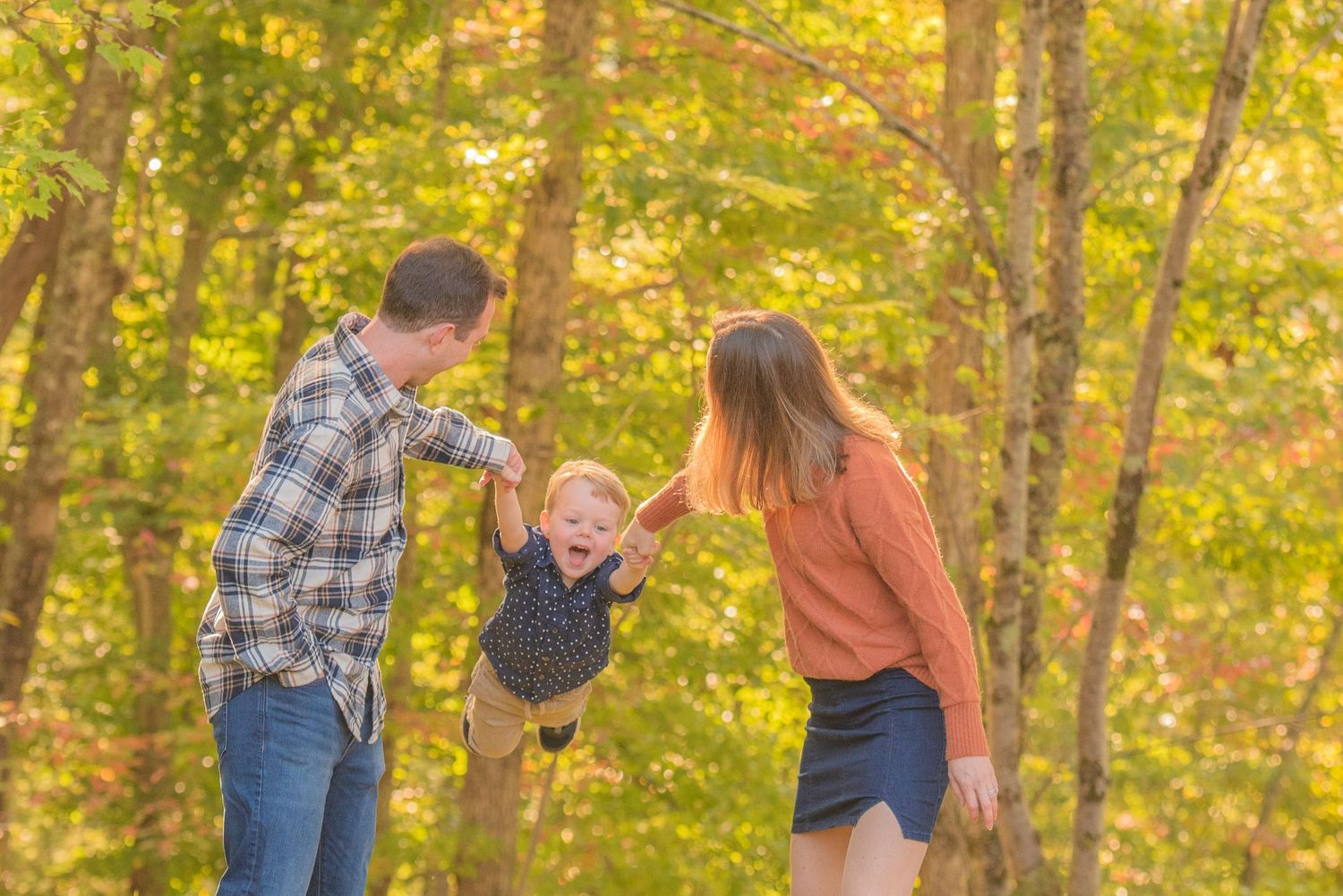 A family plays together in a sunlit autumn forest, swinging a small child between them against golden foliage in Pigeon Forge TN.