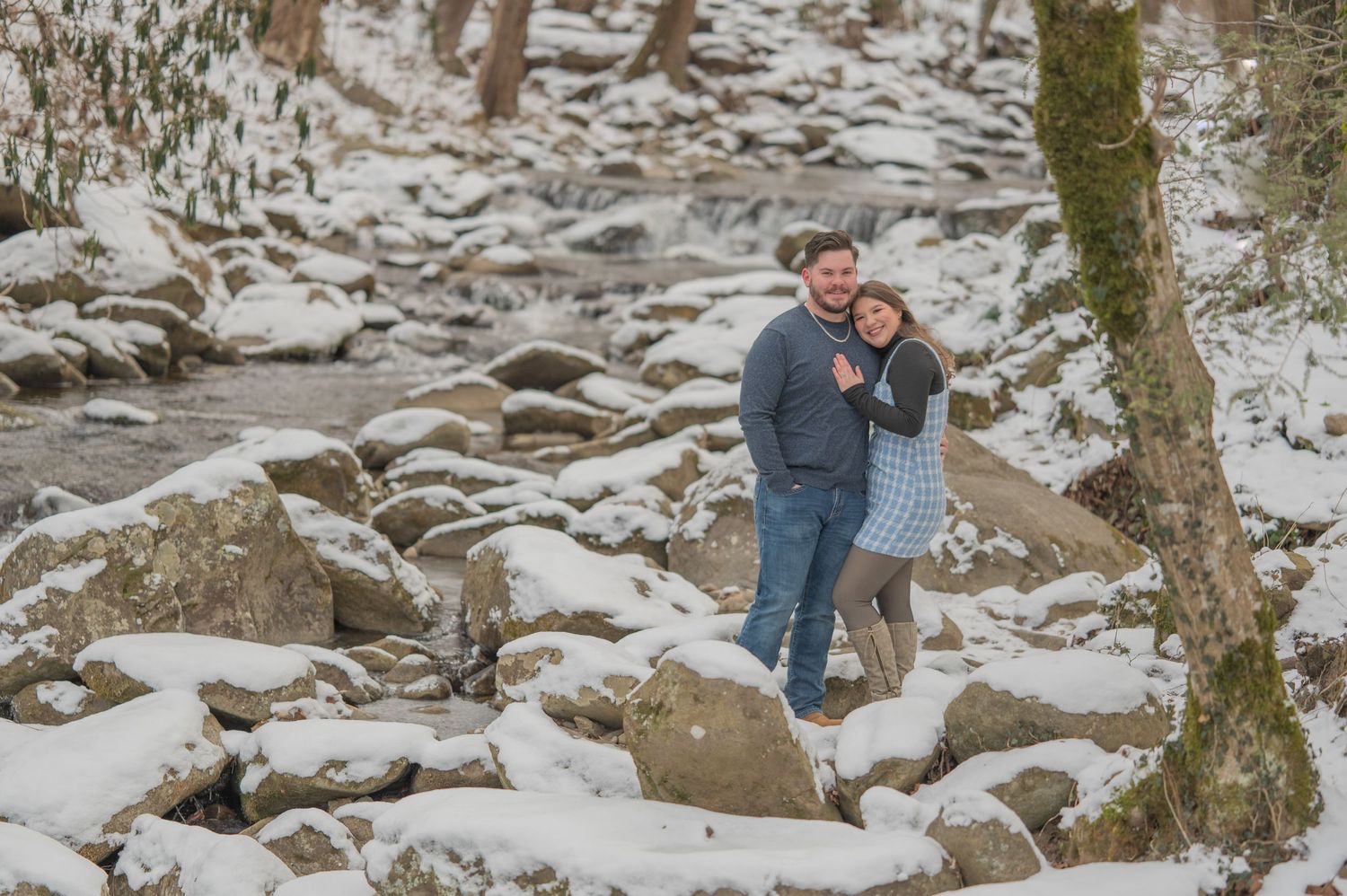 A couple stands together on snow-covered rocks near a winter stream in a romantic outdoor setting by seviervile tn engagement photographers.