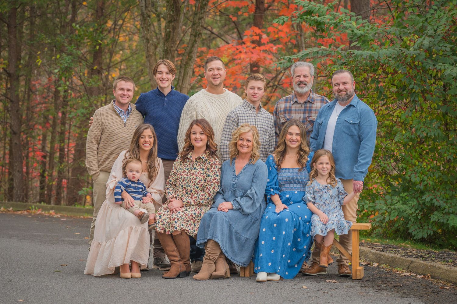 A multi-generational family group poses outdoors on a fall day with colorful autumn foliage in the background in Sevierville TN.