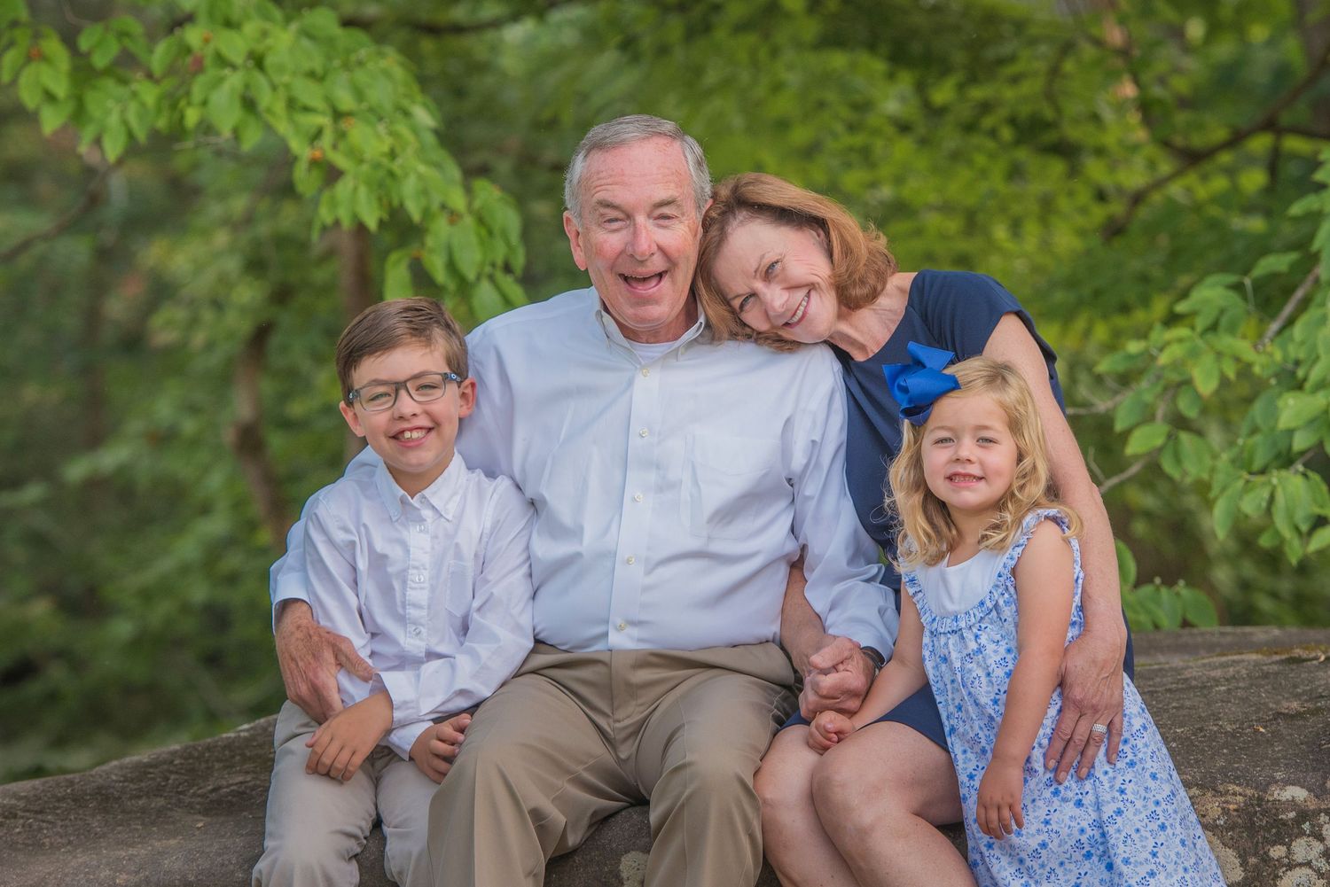 A multigenerational family group sits together outdoors on a stone wall with green foliage in the background.