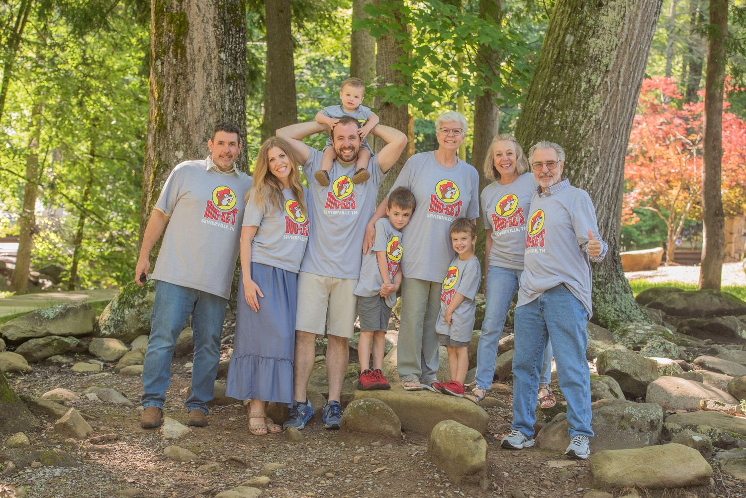 Family group wearing matching gray t-shirts with colorful logo poses together on stone path in wooded outdoor setting in Townsend TN.