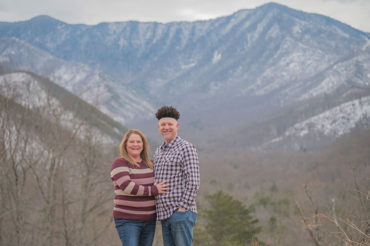 A couple stands together smiling with snow-capped mountains of the Smoky Mountains National Park in the background in Gatlinburg TN.
