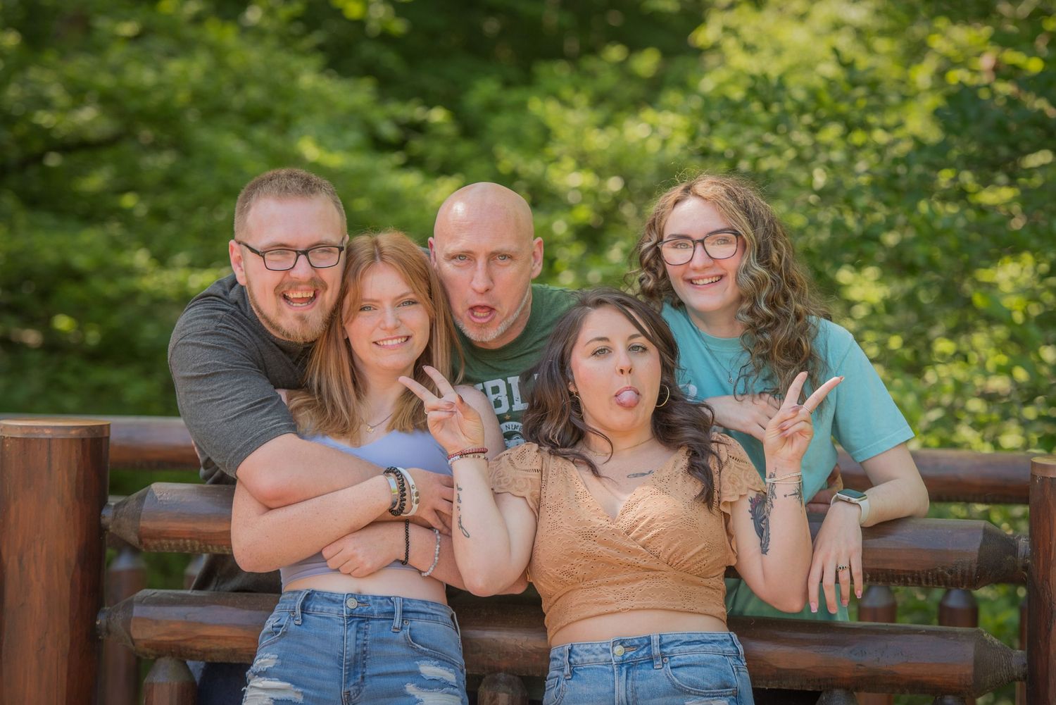 Family of five pose playfully together on a wooden bench in a lush green outdoor setting in Pigeon Forge TN.