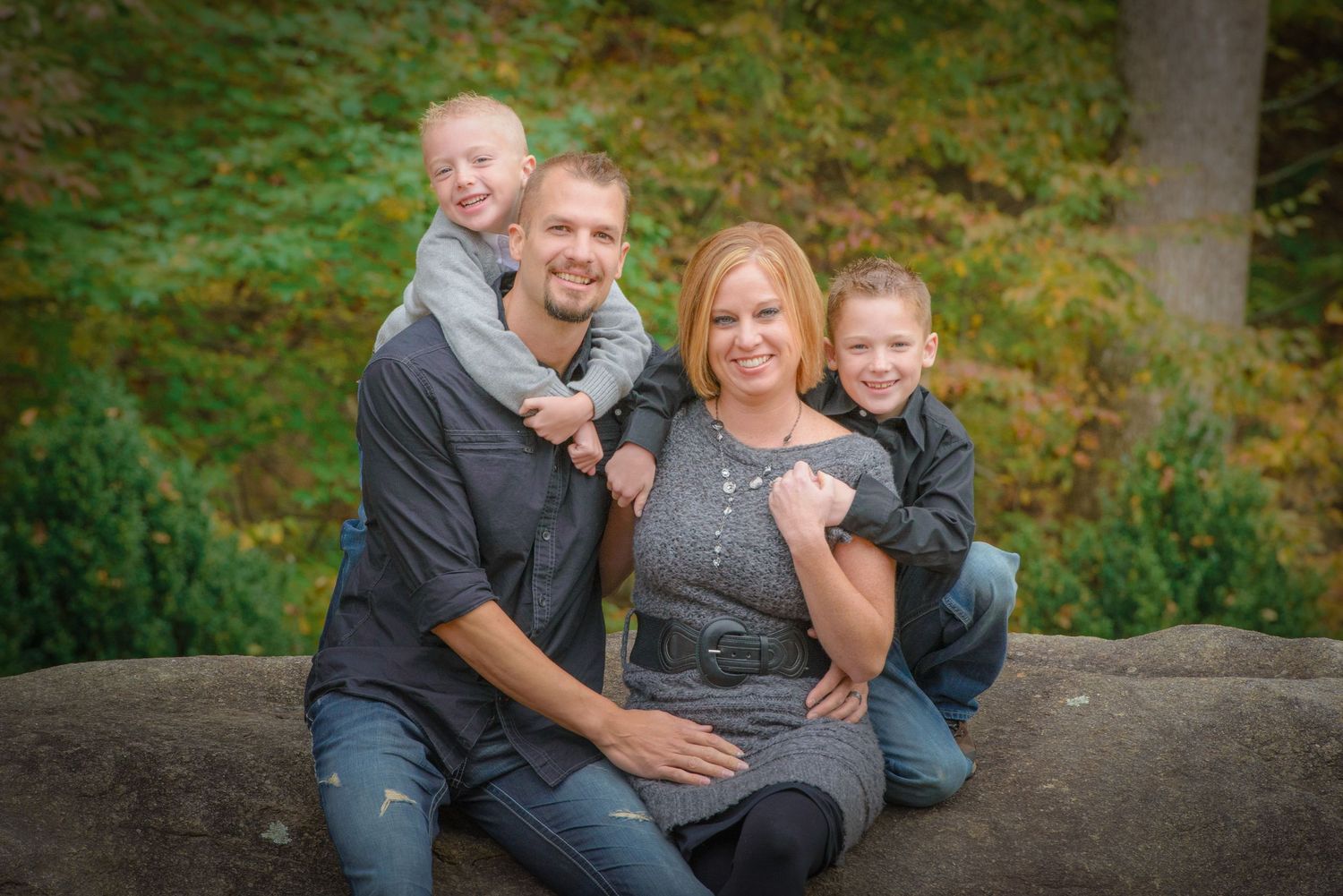A family of four sits together and smiles while posing outdoors on a rock with greenery in the background by Wears Valley Photographers.