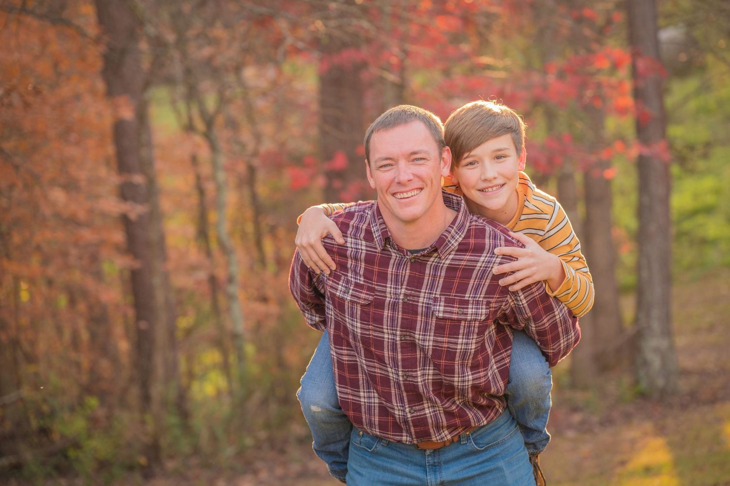 Dad and son enjoying a piggyback ride in an autumn forest with colorful fall foliage in the background by Sevierville Photographers TN.