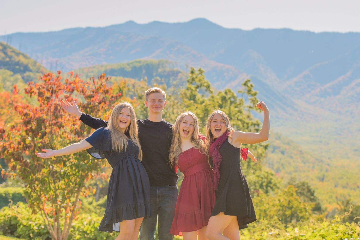 Family group poses joyfully with arms raised against backdrop of autumn foliage and mountain scenery on sunny day in Sevierville TN.
