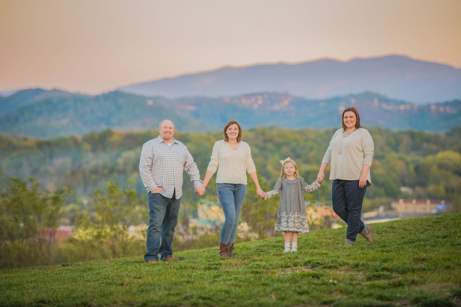 A family of four holds hands while walking on a grassy hilltop at sunset with mountains in the background.