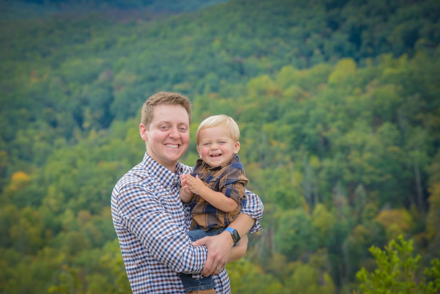 Father and son share a joyful moment while standing on a hillside with lush green forest in the background by Gatlinburg TN photographers.