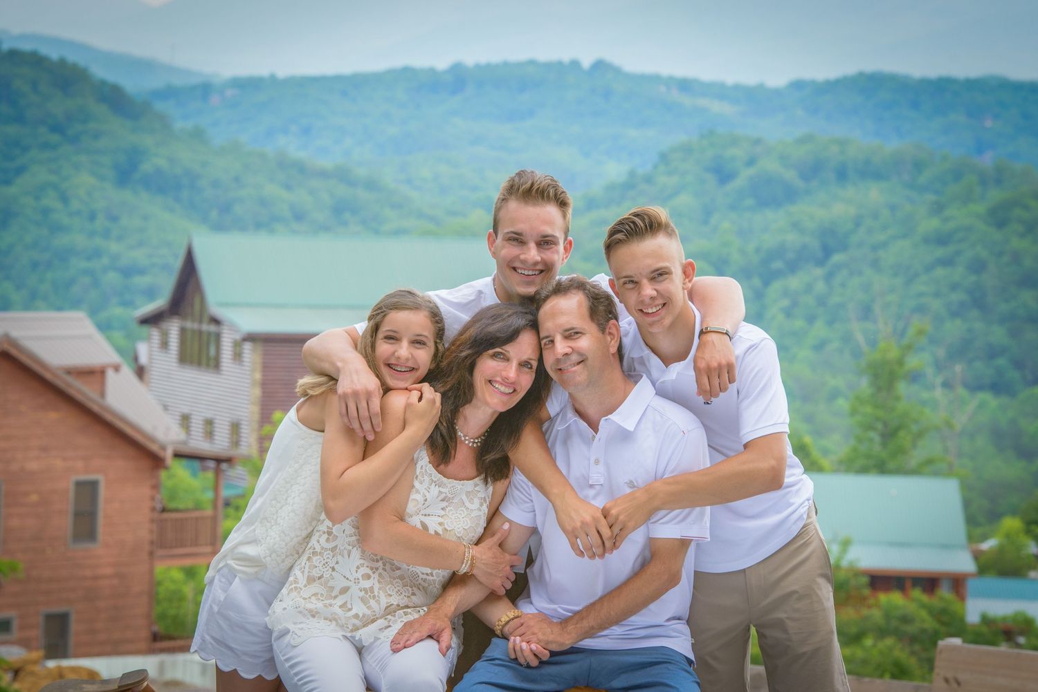 A family stands close together smiling outdoors with mountains and a church steeple visible in the background at their rental cabin.