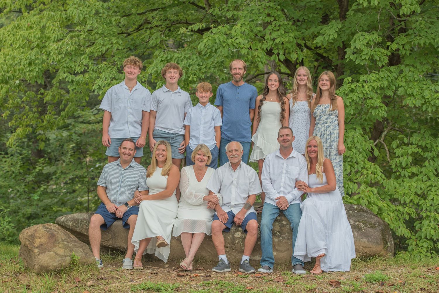 A large family group poses together outdoors on a rock with lush green trees in the background.