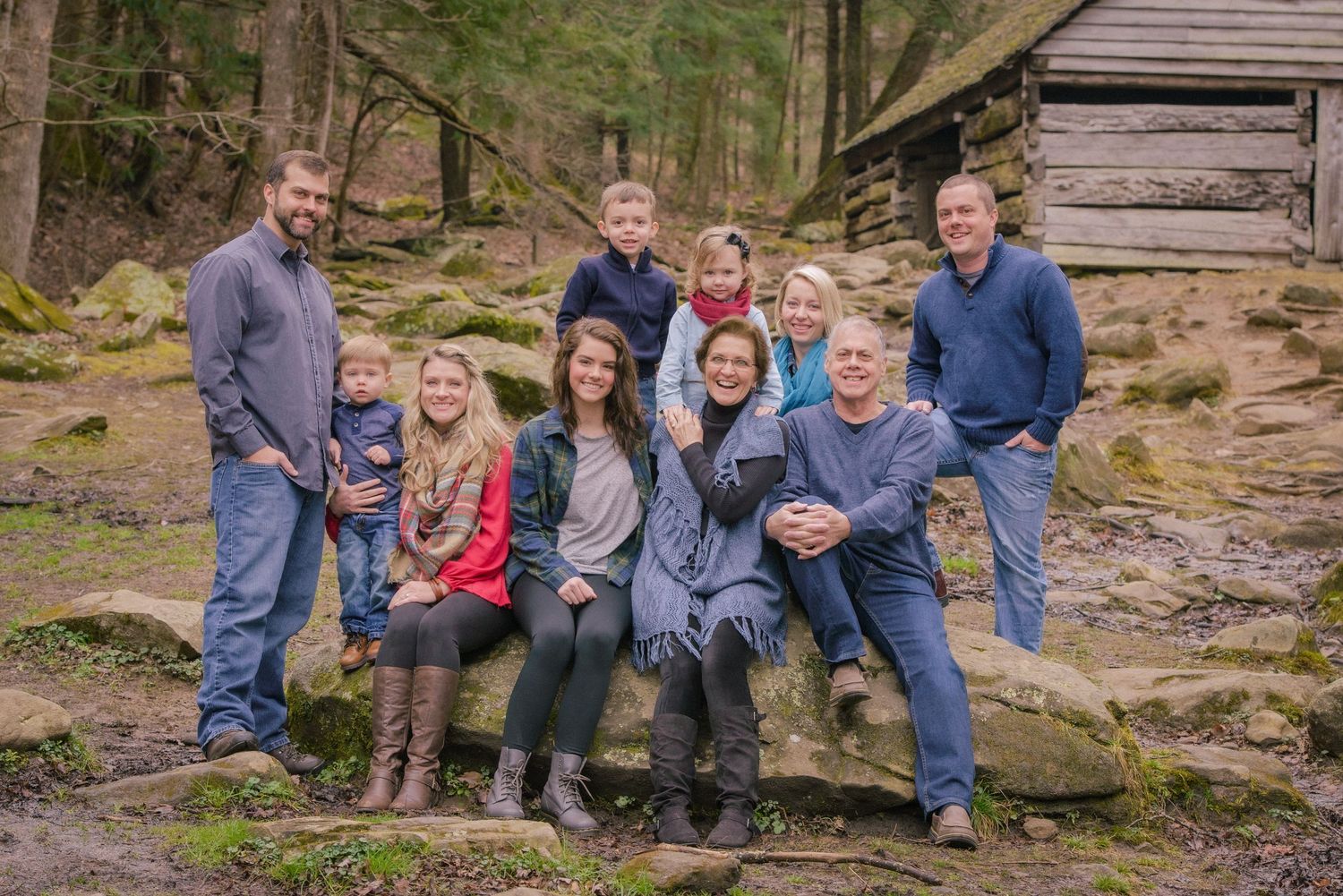 A family in denim and casual wear poses together on rocky terrain near a rustic wooden cabin in the woods Pigeon Forge TN