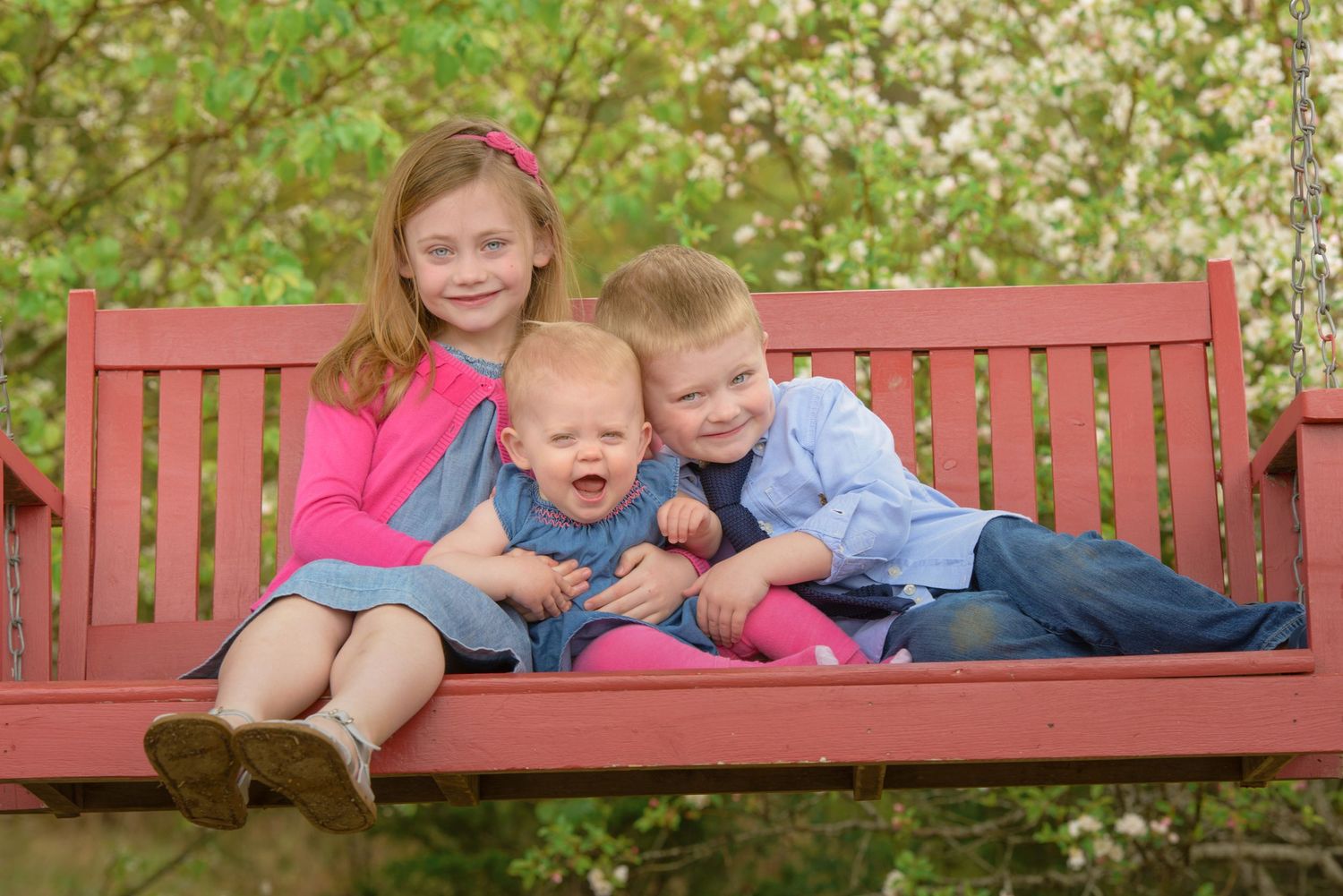 Three children sitting together and laughing on a wooden bench in a garden with blooming trees in Dandridge TN.