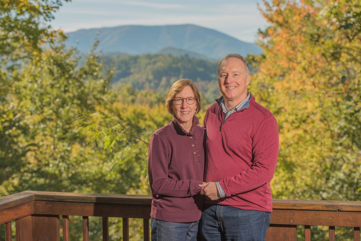 Couple in matching burgundy sweaters stand on a deck surrounded by autumn foliage and mountain views in Pigeon Forge TN.