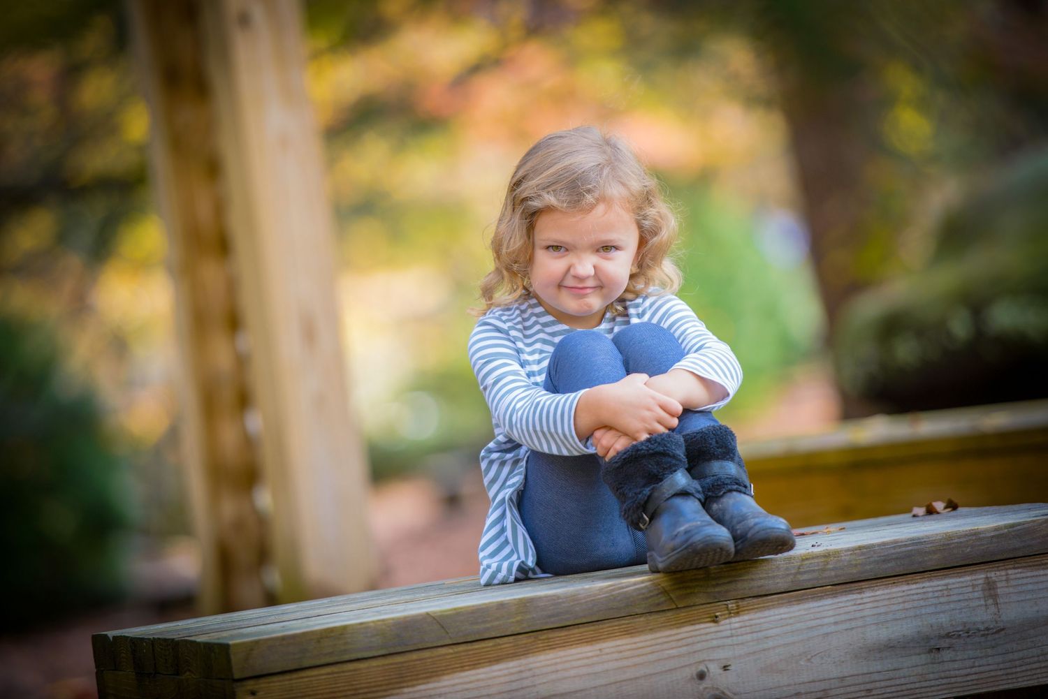 Happy child wearing blue and white striped shirt sits outdoors on wooden beam in autumn setting by Gatlinburg TN photographers.