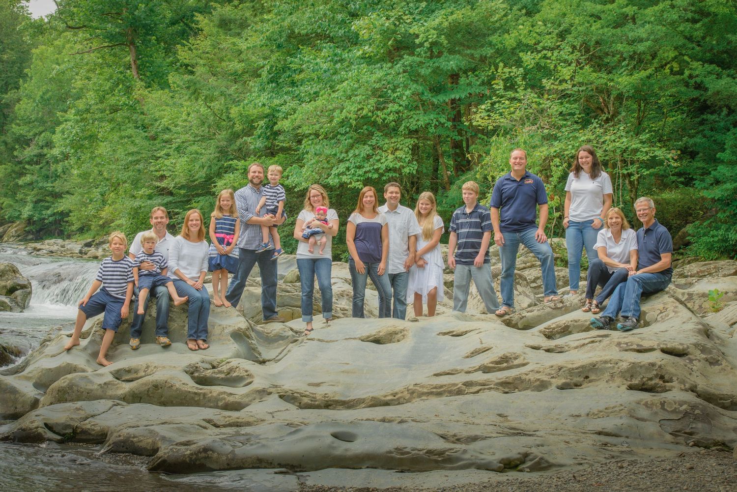 A large group stands together on a rocky riverbank surrounded by lush green trees on a sunny day.