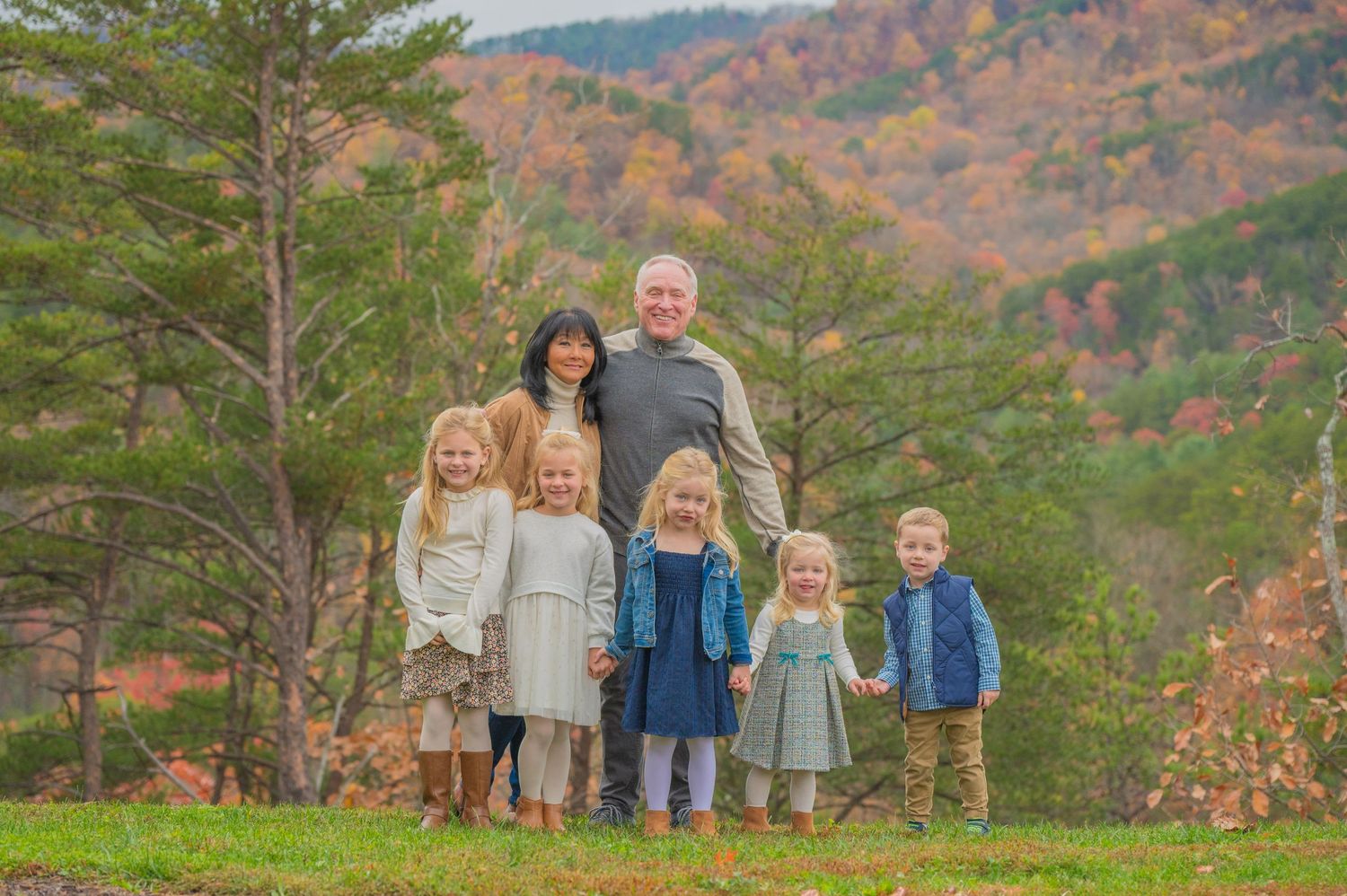A family stands together outdoors on a grassy hill with autumn mountains and evergreen trees in the background in Cosby TN.