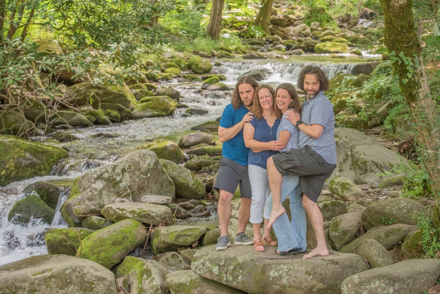 A small family stands together on mossy rocks by a flowing mountain stream in a lush green forest setting in Townsend TN.