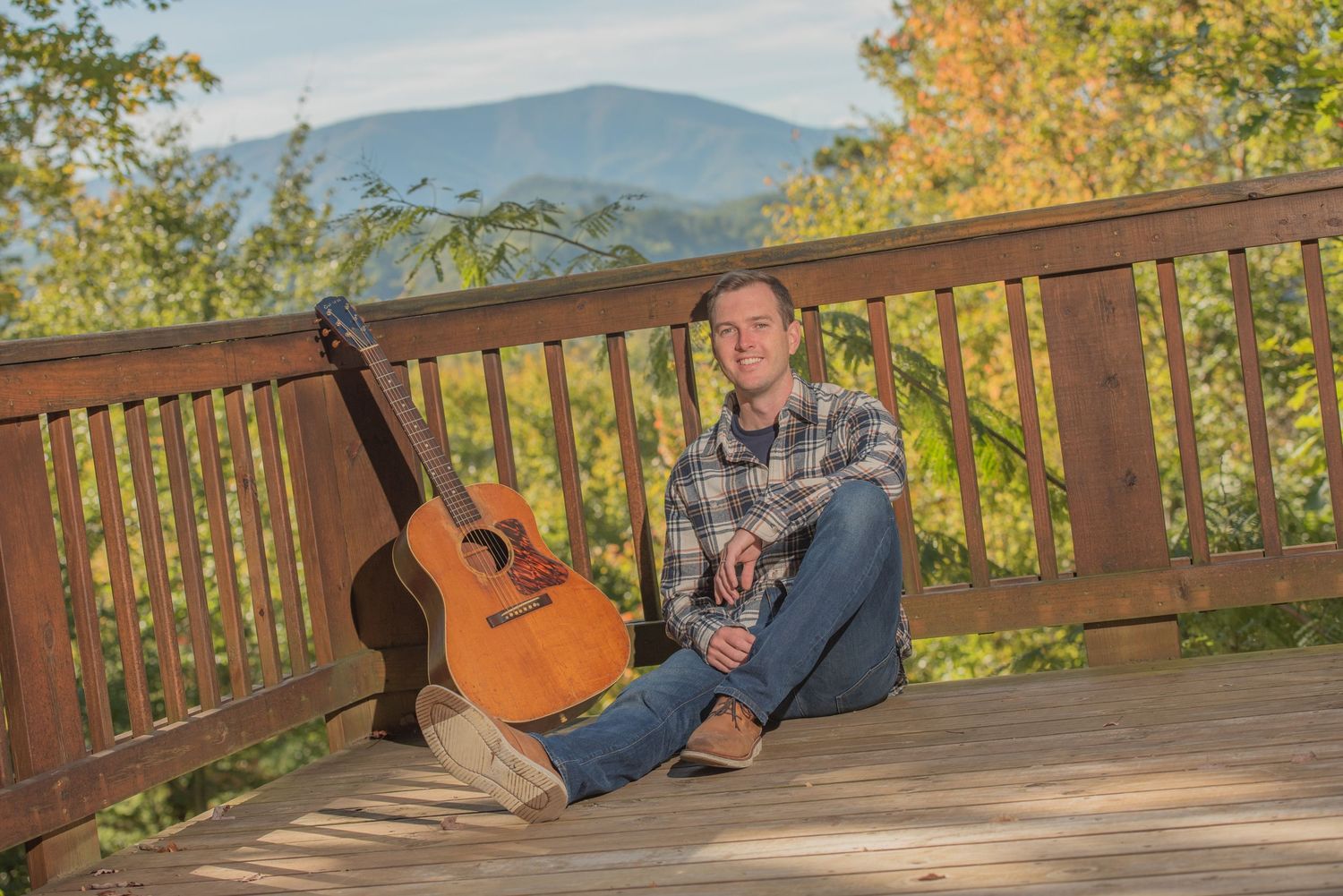 Acoustic guitar rests on wooden deck railing with autumn mountain landscape and fall foliage in background in Gatlinburg TN.
