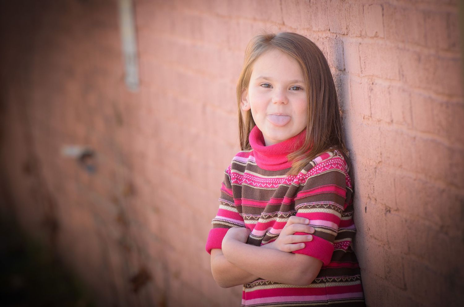 Pigeon Forge Photographer portraits of a child in a pink striped sweater stands with arms crossed against a brick wall outdoors.
