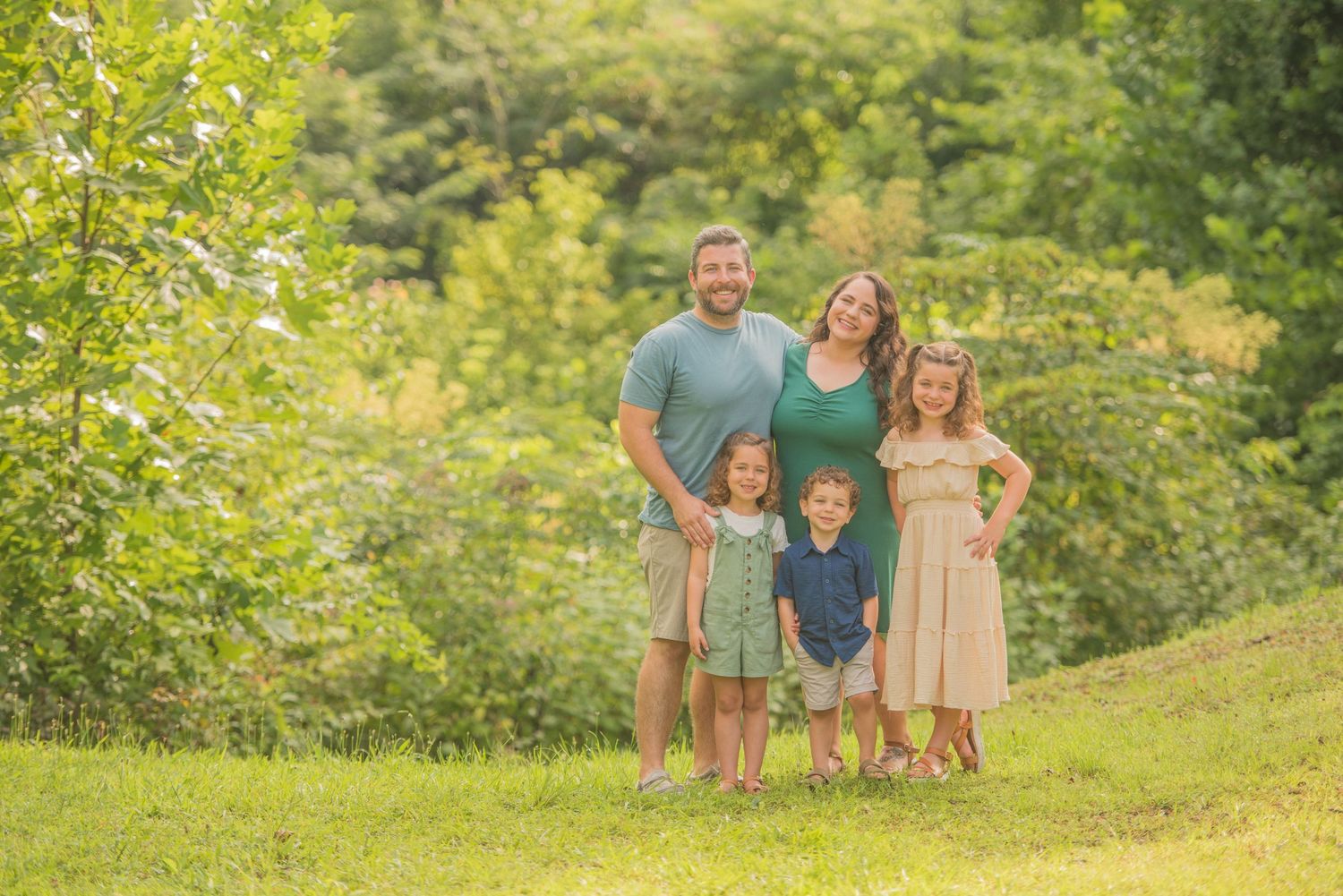 Pigeon Forge Photographers portrait of a family stands together on a grassy hillside surrounded by lush green foliage on a sunny summer day.