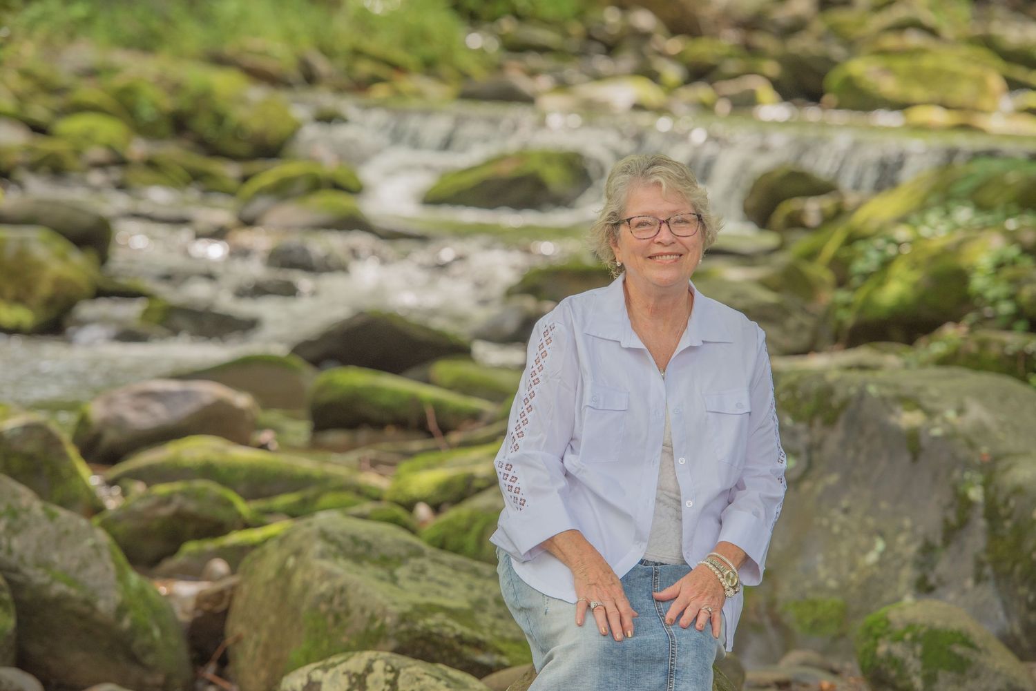 A grandma in a light blue button-down shirt and jeans sits on moss-covered rocks by a flowing stream by Gatlinburg Photographers.