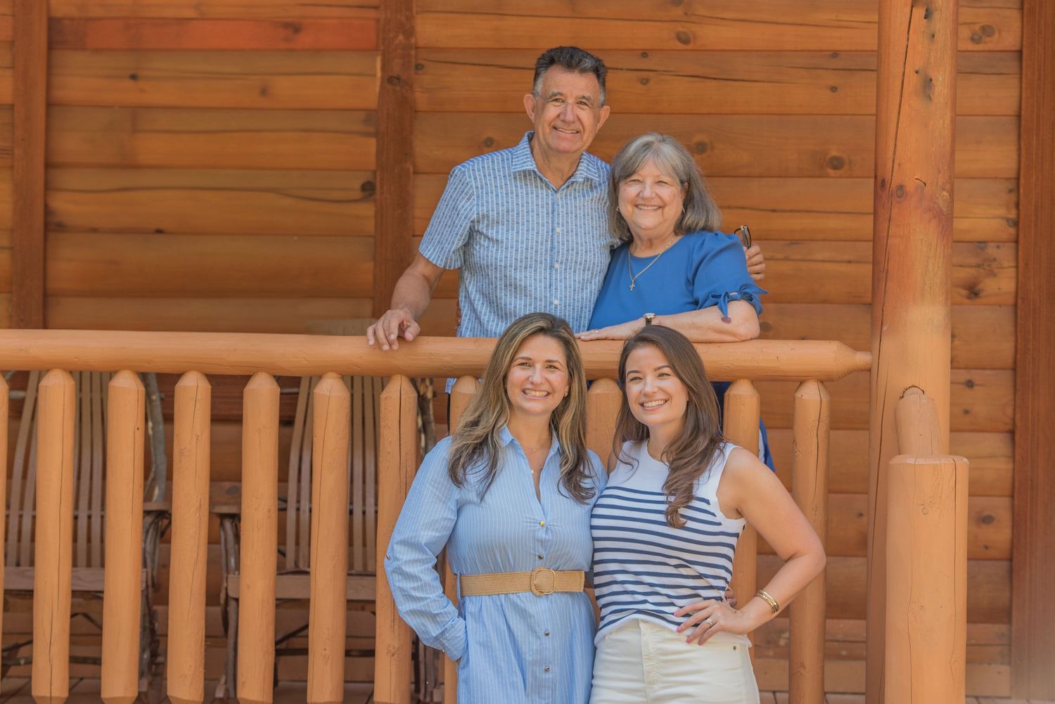 A family of four poses on a rustic wooden cabin porch with log railing on a sunny day Pigeon Forge TN.
