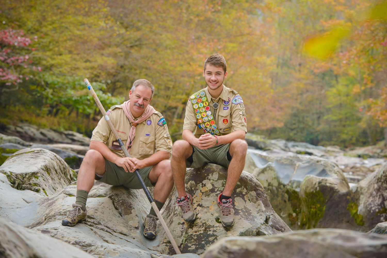 Dad and son scouts in uniform sit on rocky terrain by a stream surrounded by autumn foliage in the background in the Great Smoky Mountains.