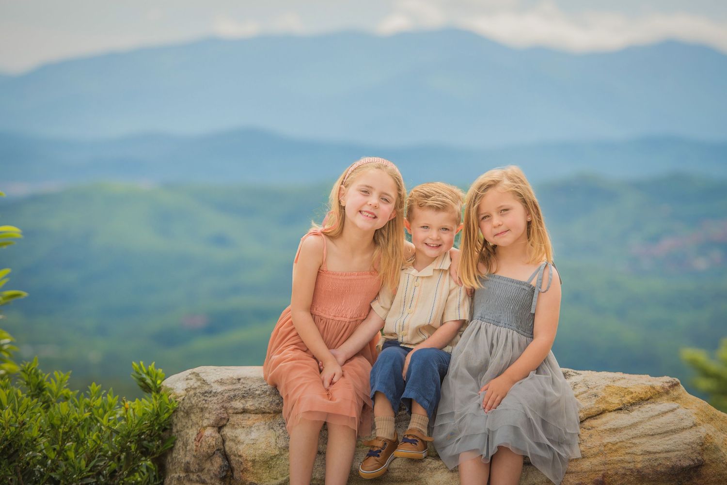 Three children sit together on a rock overlook with misty blue mountains visible in the background.