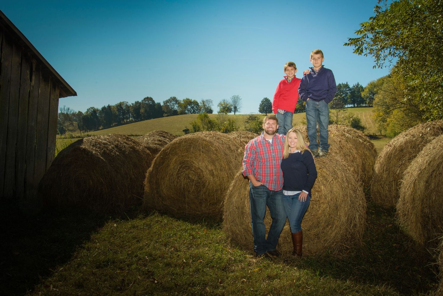 A farm family stands atop large round hay bales in a sunny field beside a rustic barn on a clear autumn day in Sevierville TN.