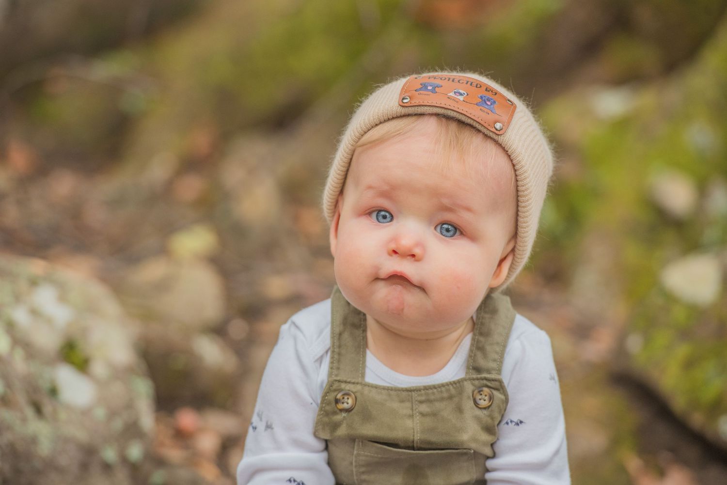 Gatlinburg Tn Photographer portrait of Toddler wearing brown overalls and beige cap sits outdoors against natural rocky background.