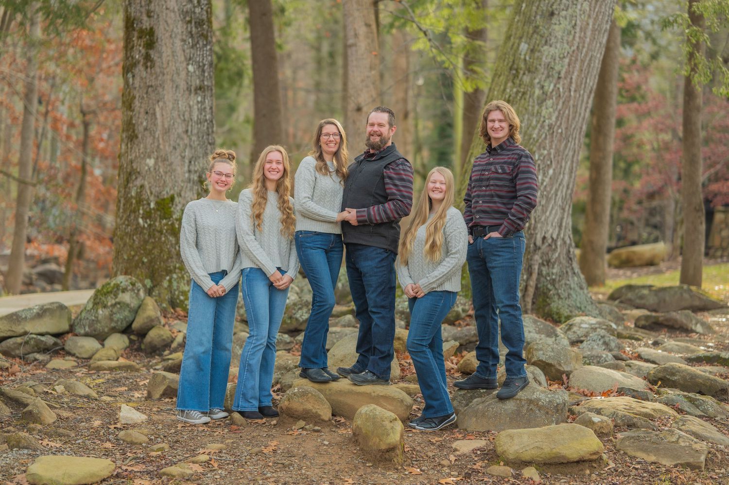 Family of six poses outdoors in matching denim and sweaters on rocky wooded terrain during autumn by Gatlinburg TN Photographers.