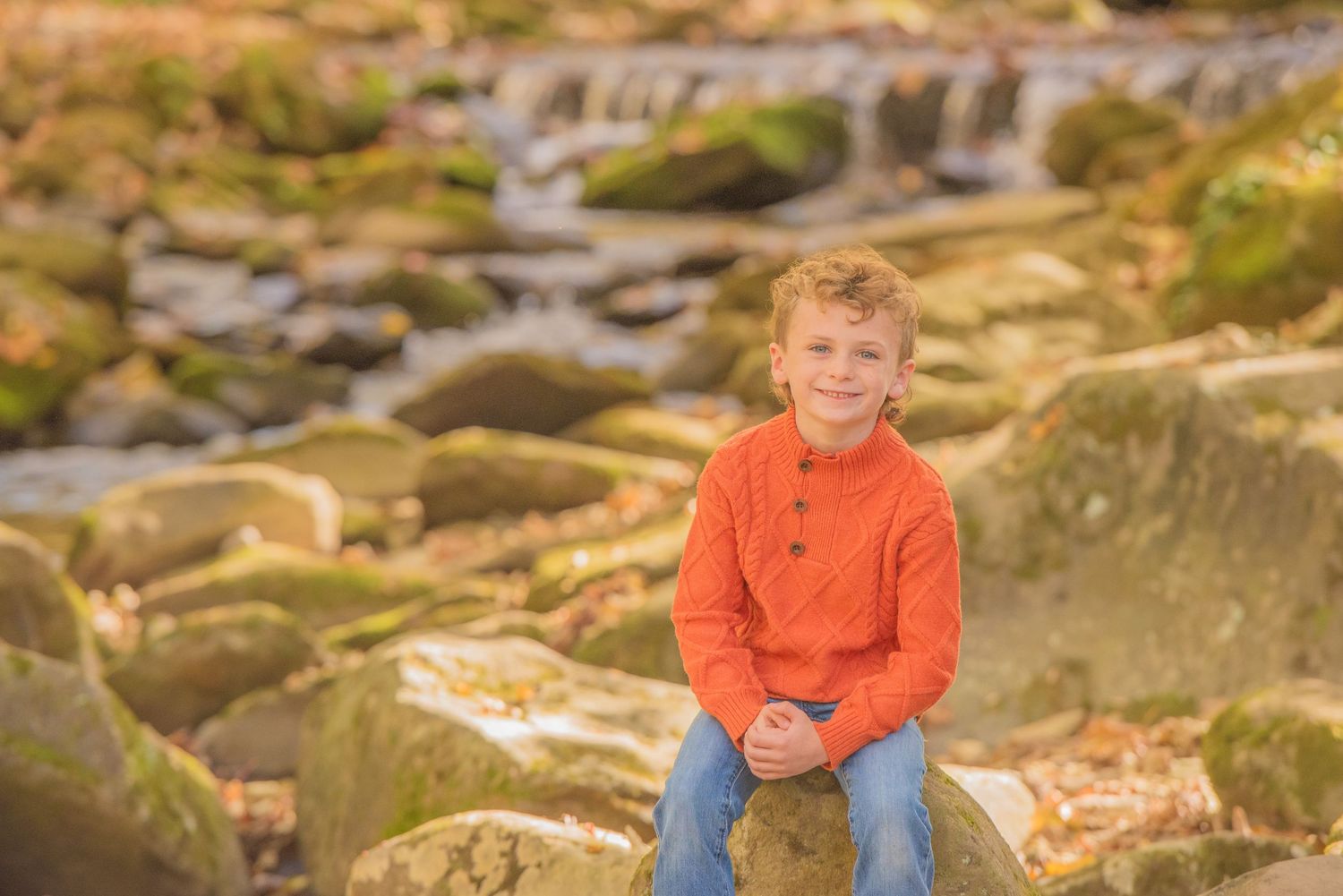 A child in a bright orange sweater sits relaxed on mossy rocks near a cascading waterfall in warm lighting in Gatlinburg TN.