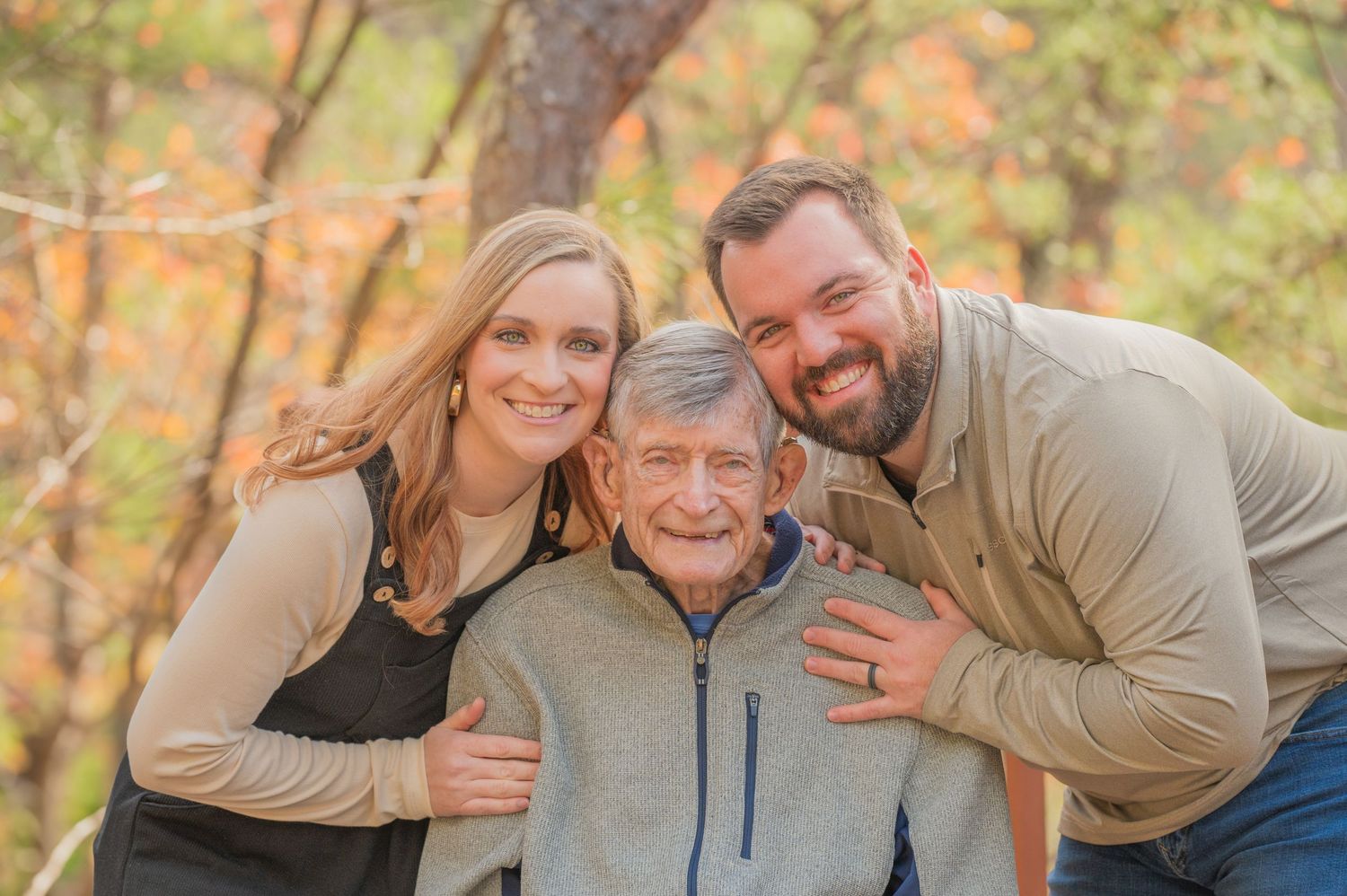 Two grown grandkids taking a portrait with grandpa outdoors in autumn with fall foliage and trees in the background in Sevierville Cabin.