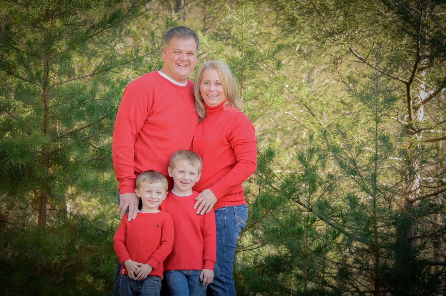 A family of three wearing matching red sweaters poses together in front of green trees for a holiday portrait by Sevierville Photographers.